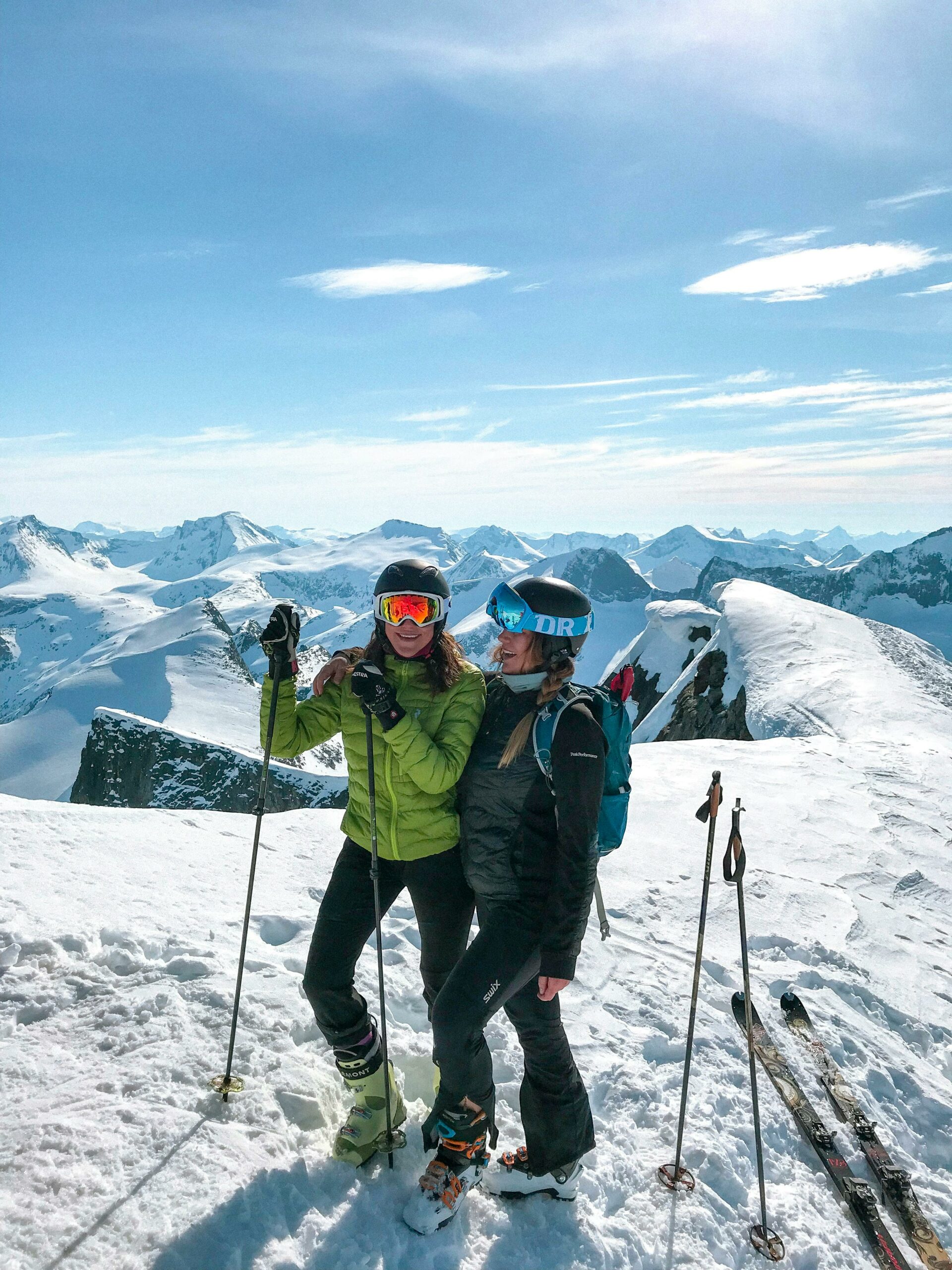 Two skiers enjoy a sunny winter day on a snowy mountain peak with stunning panoramic views.