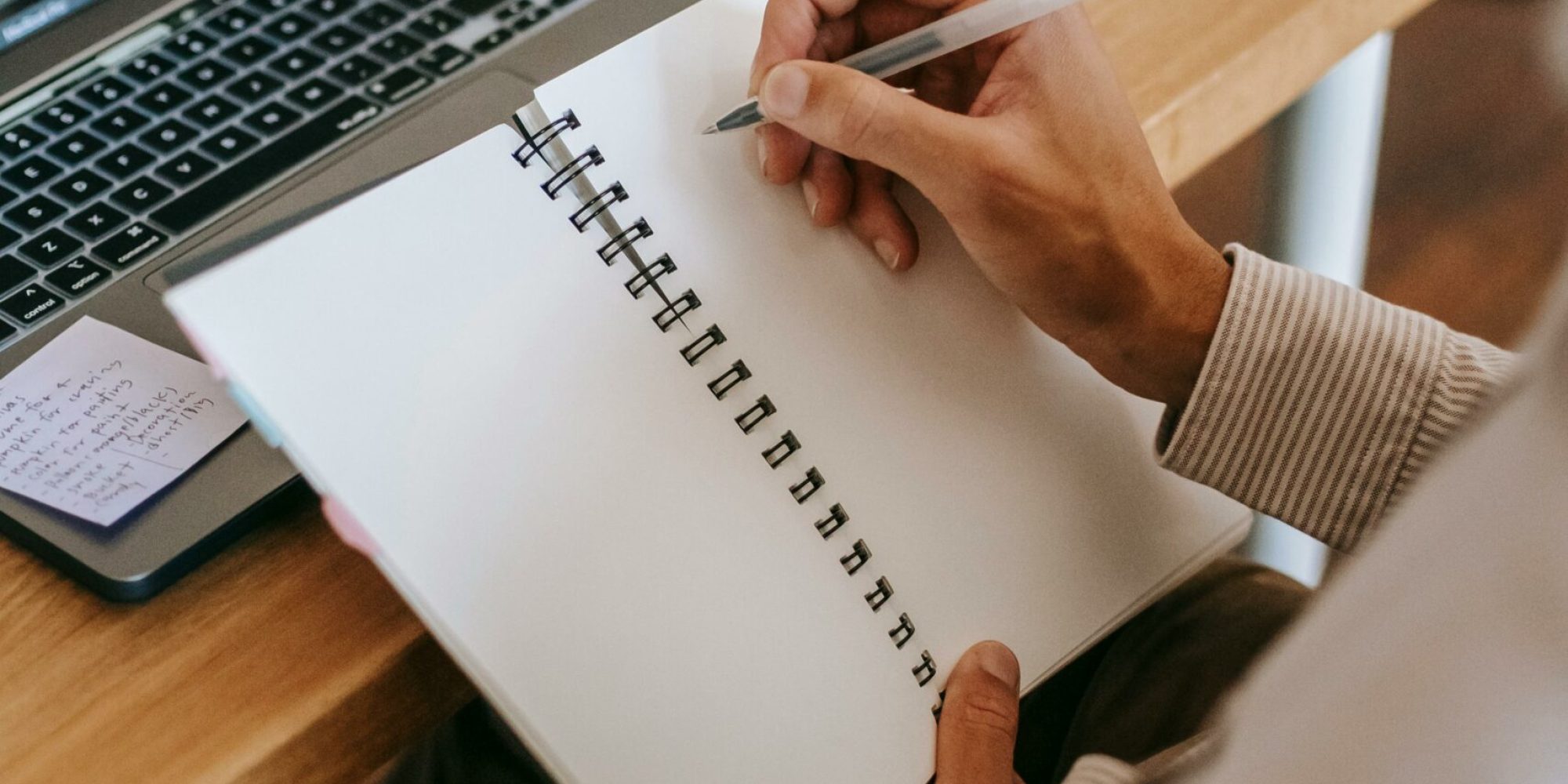 Man writing in notebook with laptop and coffee in home office setting.