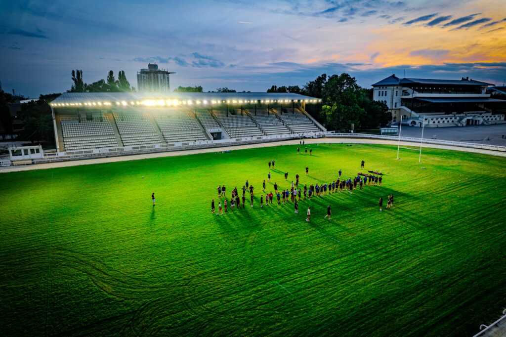 Aerial shot of a group practicing rugby under stadium lights at sunset, creating a vibrant and dynamic atmosphere.